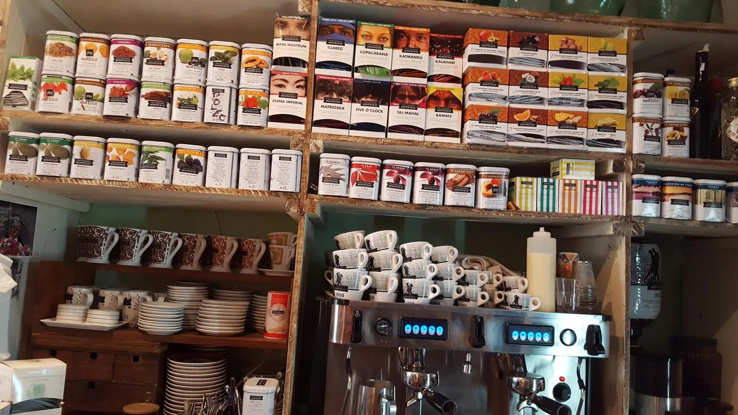 Estantes de tienda con una variedad de tés y especias enlatados, junto a una máquina de café y tazas apiladas. Ideal para amantes del té y el café.