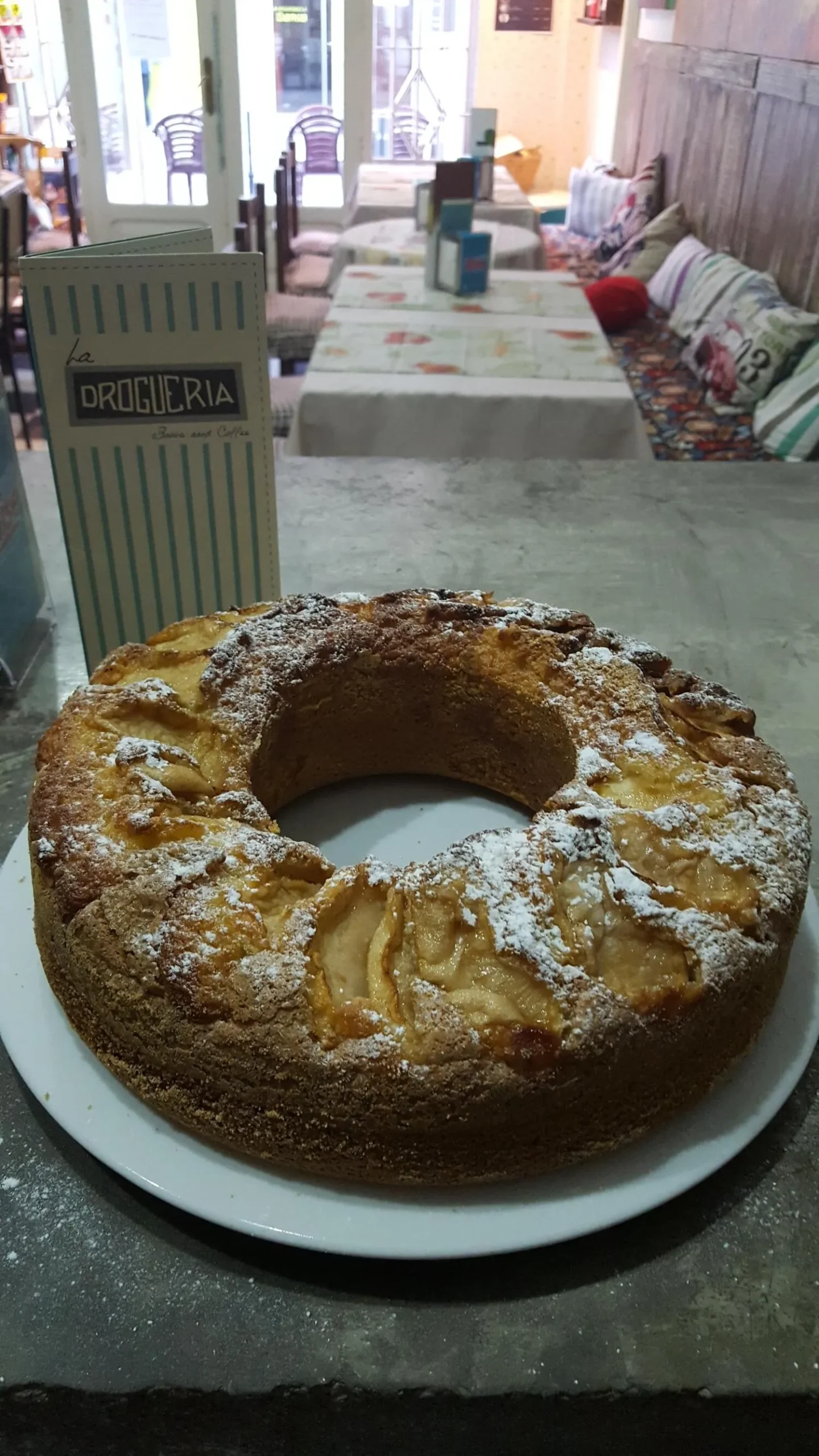 Tarta de manzana recién horneada, espolvoreada con azúcar glas, presentada en un plato blanco. En el fondo, se puede ver el menú de "La Droguería" y un ambiente acogedor de cafetería.