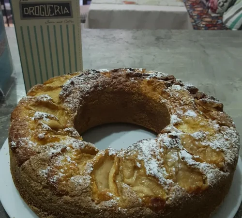 Tarta de manzana recién horneada, espolvoreada con azúcar glas, presentada en un plato blanco. En el fondo, se puede ver el menú de "La Droguería" y un ambiente acogedor de cafetería.